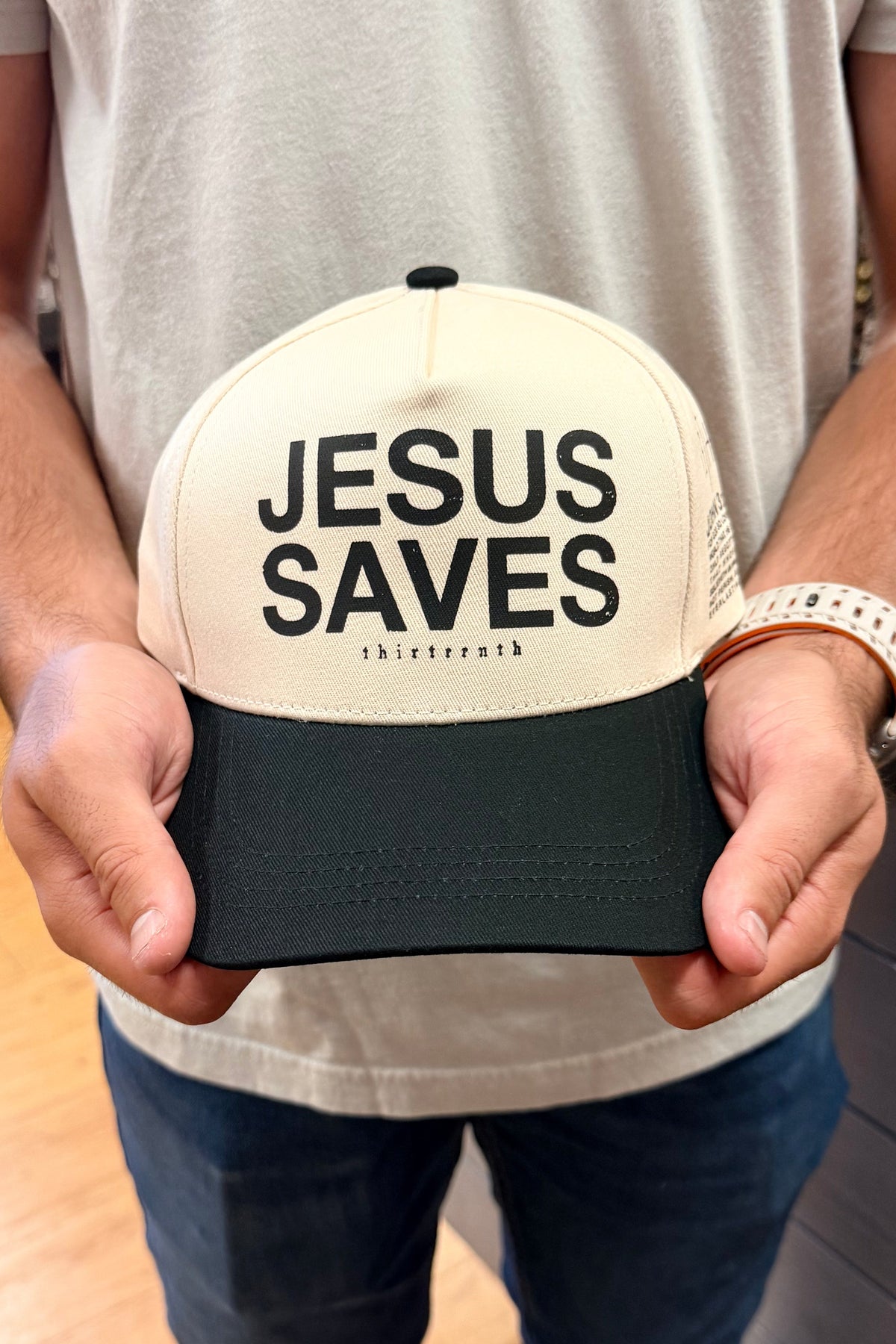 A man in a beige t-shirt and jeans holds a cream and dark green trucker hat with the text 'JESUS SAVES' printed on the front. The photo is taken inside a retail store.