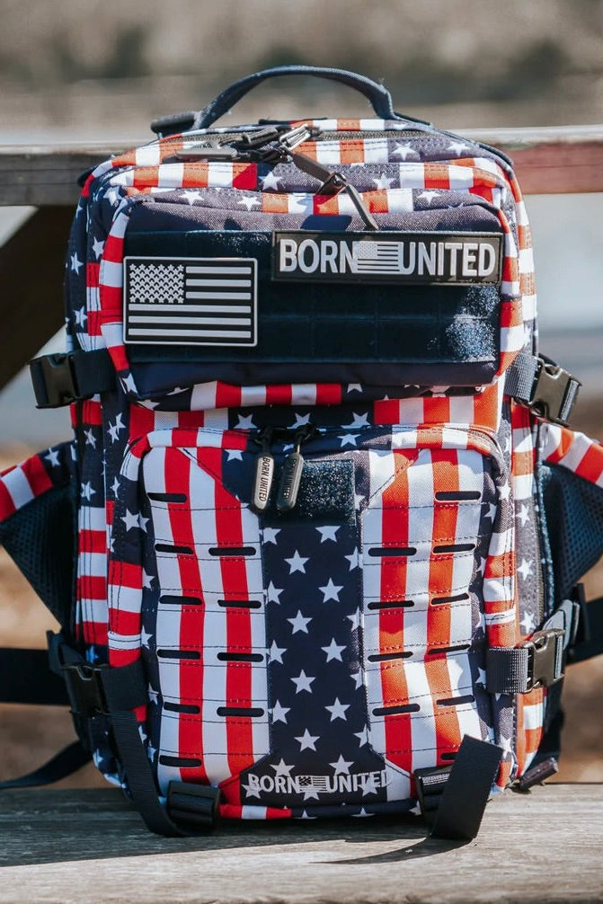The Premium Tac-Pac Patch Bundle by Born United Private Label is a backpack with an American flag pattern, stars, red, white and blue stripes, plus BORN UNITED and American flag patches displayed on a wooden outdoor bench.