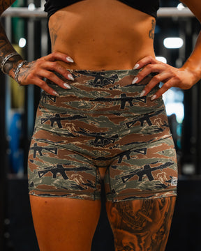 A person stands hands on hips in Tango Charlie Apparels Womens High-Waisted Seamless Front Compression Training Shorts | Snipes & Stripes and a black crop top, showing tattoos on arms and leg. The gym setting highlights these compression shorts.