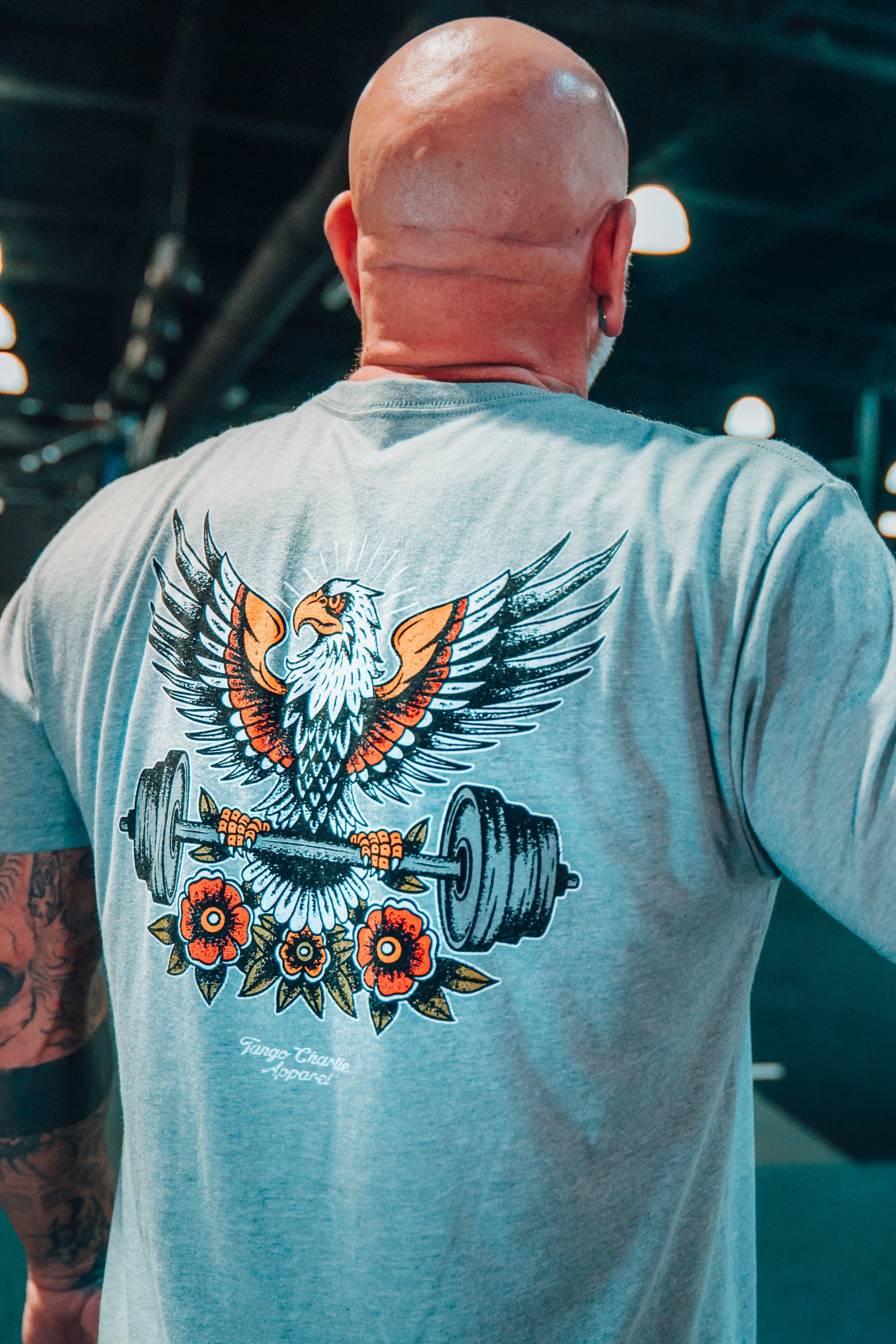 A bald, tattooed man wears Tango Charlie Apparel's "Old School Never Gets Old" gray tee with an illustrated eagle gripping weight plates and red flowers, set in a classic weightlifting gym.