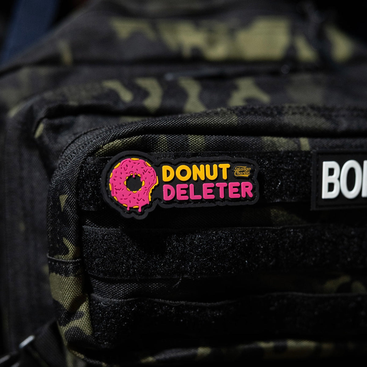 The Donut Deleter - Patch by Tango Charlie Apparel is a 3-inch 3D PVC patch with a pink frosted donut and "DONUT DELETER" text, Velcro-attached to the black front pocket of a camo backpack.