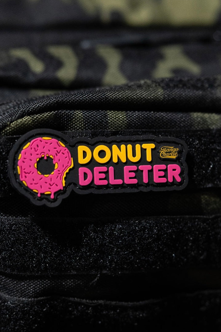 The Donut Deleter - Patch by Tango Charlie Apparel is a 3-inch 3D PVC patch with a pink frosted donut and "DONUT DELETER" text, Velcro-attached to the black front pocket of a camo backpack.