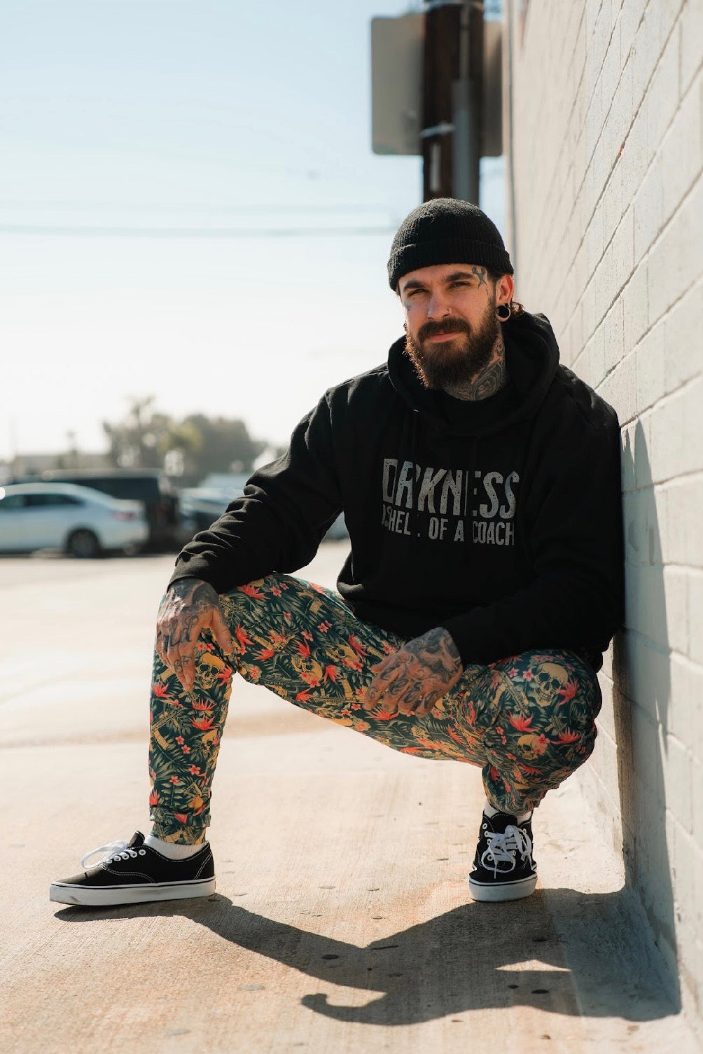A tattooed man with a beard and black beanie squats by a sunlit brick wall, wearing Tango Charlie Apparel's Men's Classic Premium Joggers | Aloha AR, a black hoodie, and black sneakers. Cars and trees appear in the background.