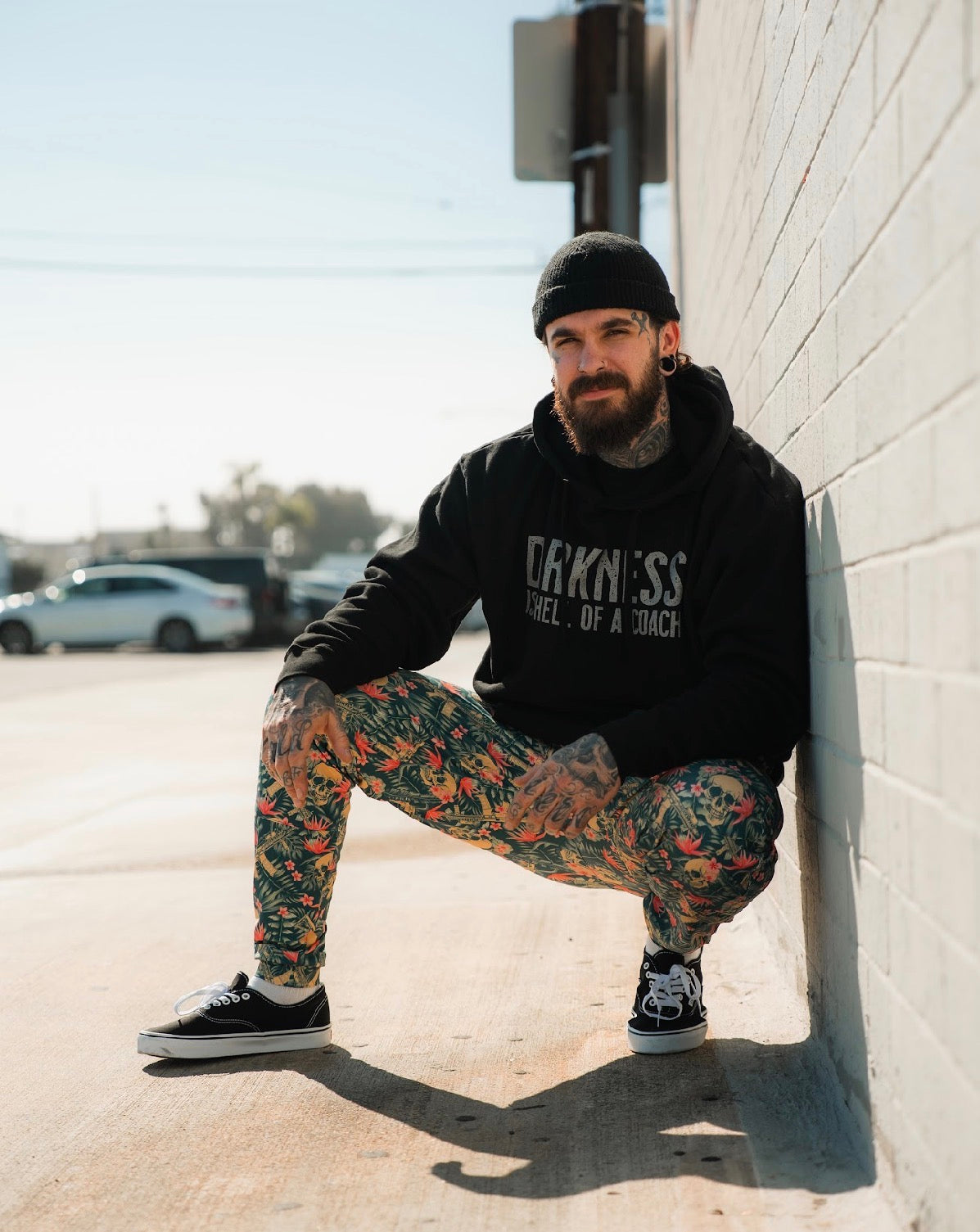 A tattooed man with a beard and black beanie squats by a sunlit brick wall, wearing Tango Charlie Apparel's Men's Classic Premium Joggers | Aloha AR, a black hoodie, and black sneakers. Cars and trees appear in the background.