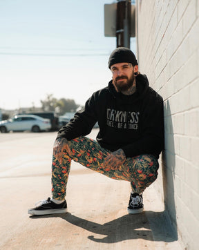 A tattooed man with a beard and black beanie squats by a sunlit brick wall, wearing Tango Charlie Apparel's Men's Classic Premium Joggers | Aloha AR, a black hoodie, and black sneakers. Cars and trees appear in the background.