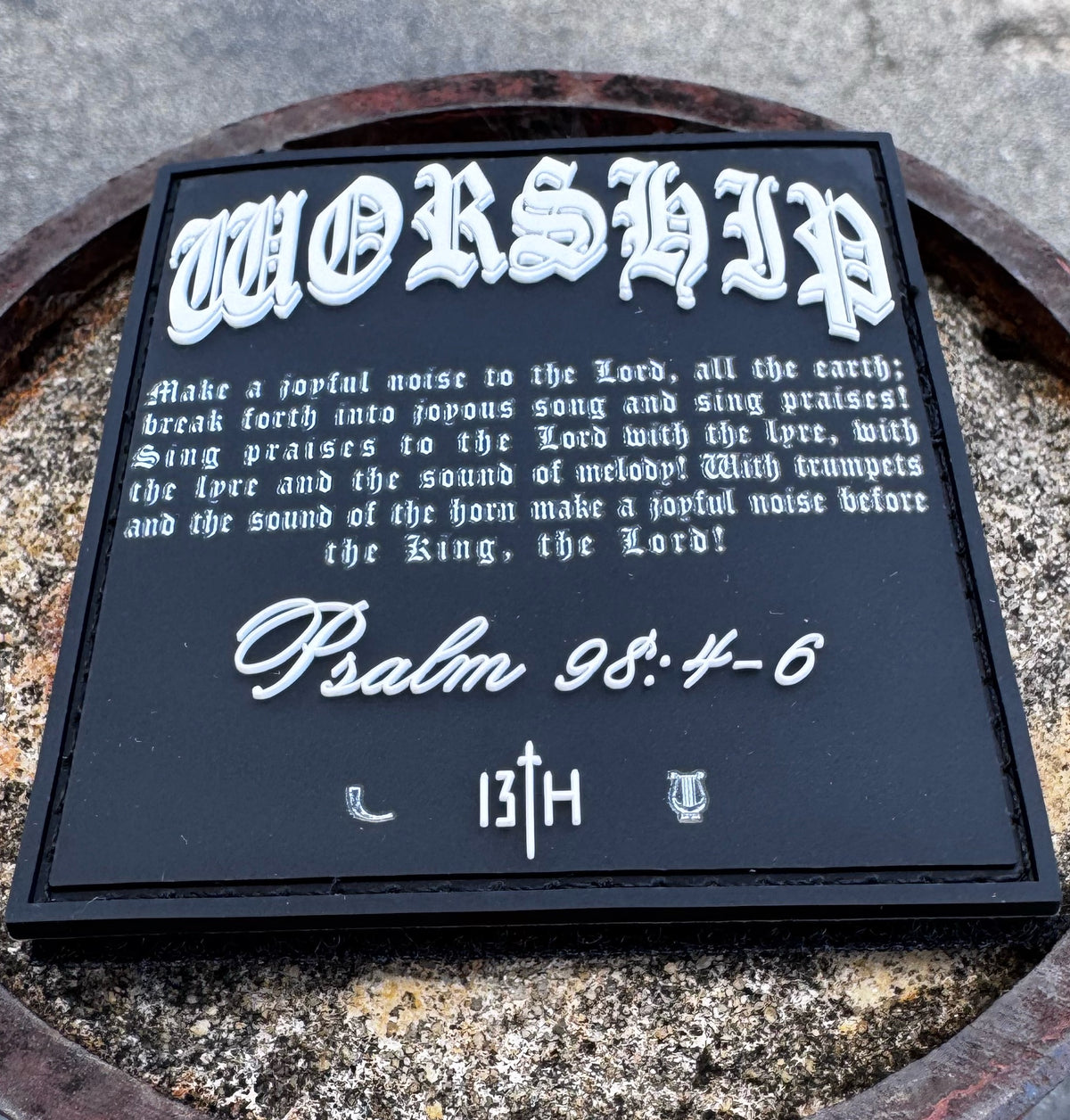 The Worship PVC Patch by 13th features Psalm 98:4-6 in ornate white text on black, with 13TH and two small symbols at the bottom, shown resting on a stone surface.