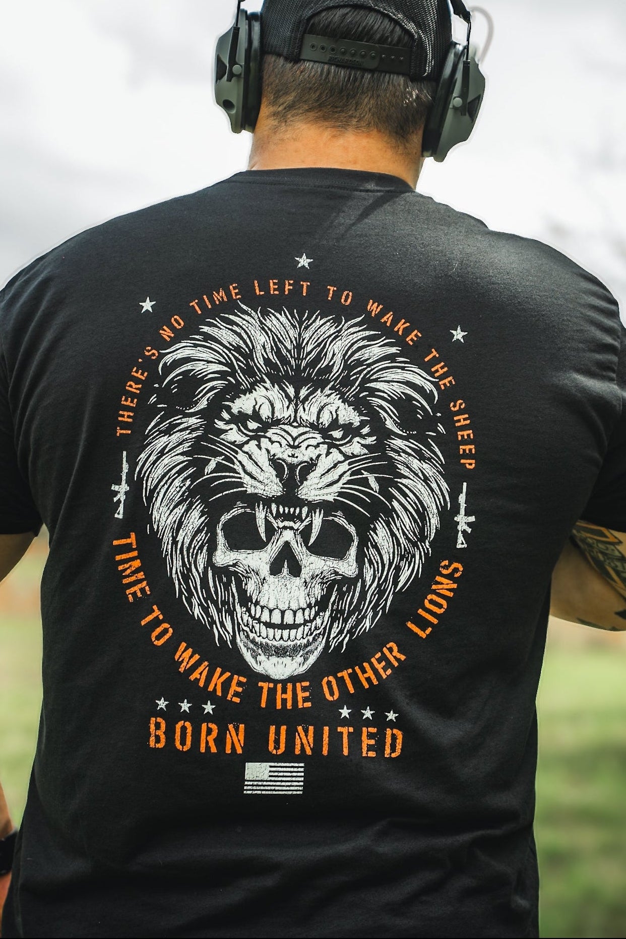 Standing outdoors, a person wears headphones and a black T-shirt featuring a lion and skull design. The shirt reads: No Time Left to Wake the Sheep, Time to Wake the Other Lions, from Born United Private Labels Waking Lions line.