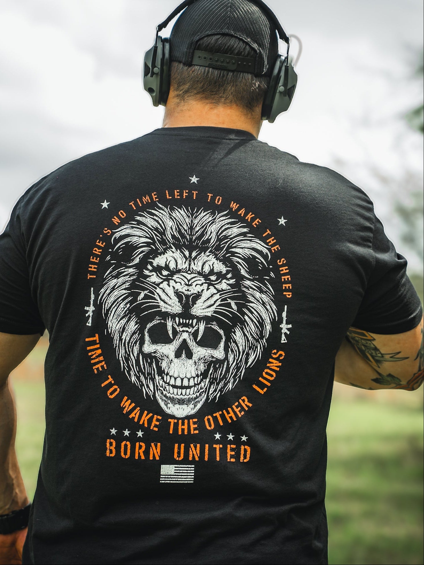 Standing outdoors, a person wears headphones and a black T-shirt featuring a lion and skull design. The shirt reads: No Time Left to Wake the Sheep, Time to Wake the Other Lions, from Born United Private Labels Waking Lions line.