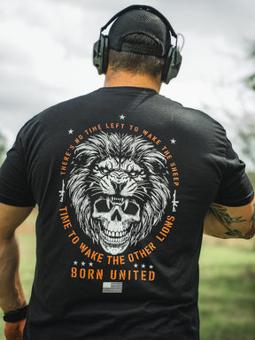 Standing outdoors, a person wears headphones and a black T-shirt featuring a lion and skull design. The shirt reads: No Time Left to Wake the Sheep, Time to Wake the Other Lions, from Born United Private Labels Waking Lions line.