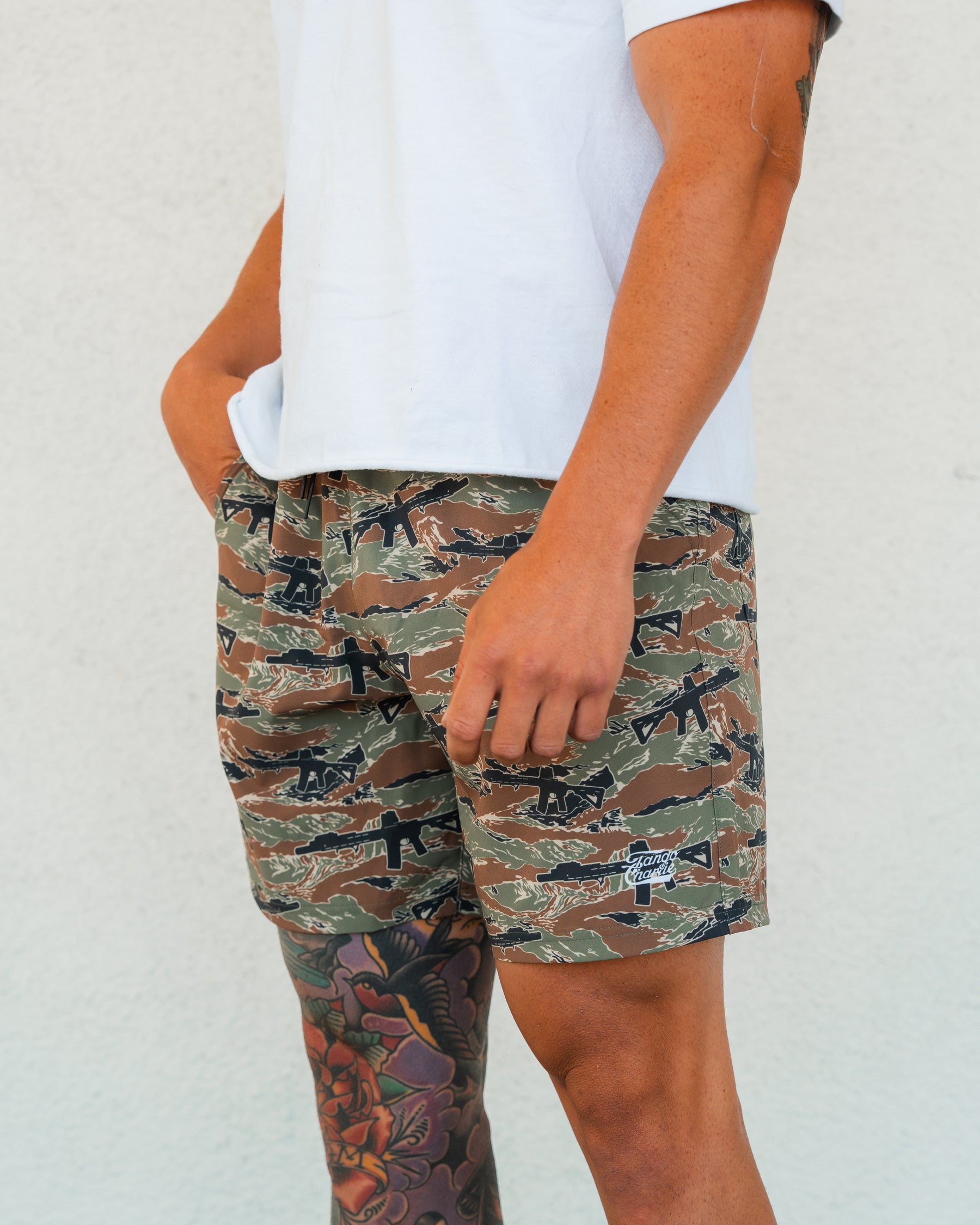 A person in a white T-shirt and Tango Charlie Apparel’s Men’s Performance Training Shorts | 5.5"- Snipes & Stripes stands against a light wall, one hand in their pocket, with colorful leg tattoos showing below the shorts.