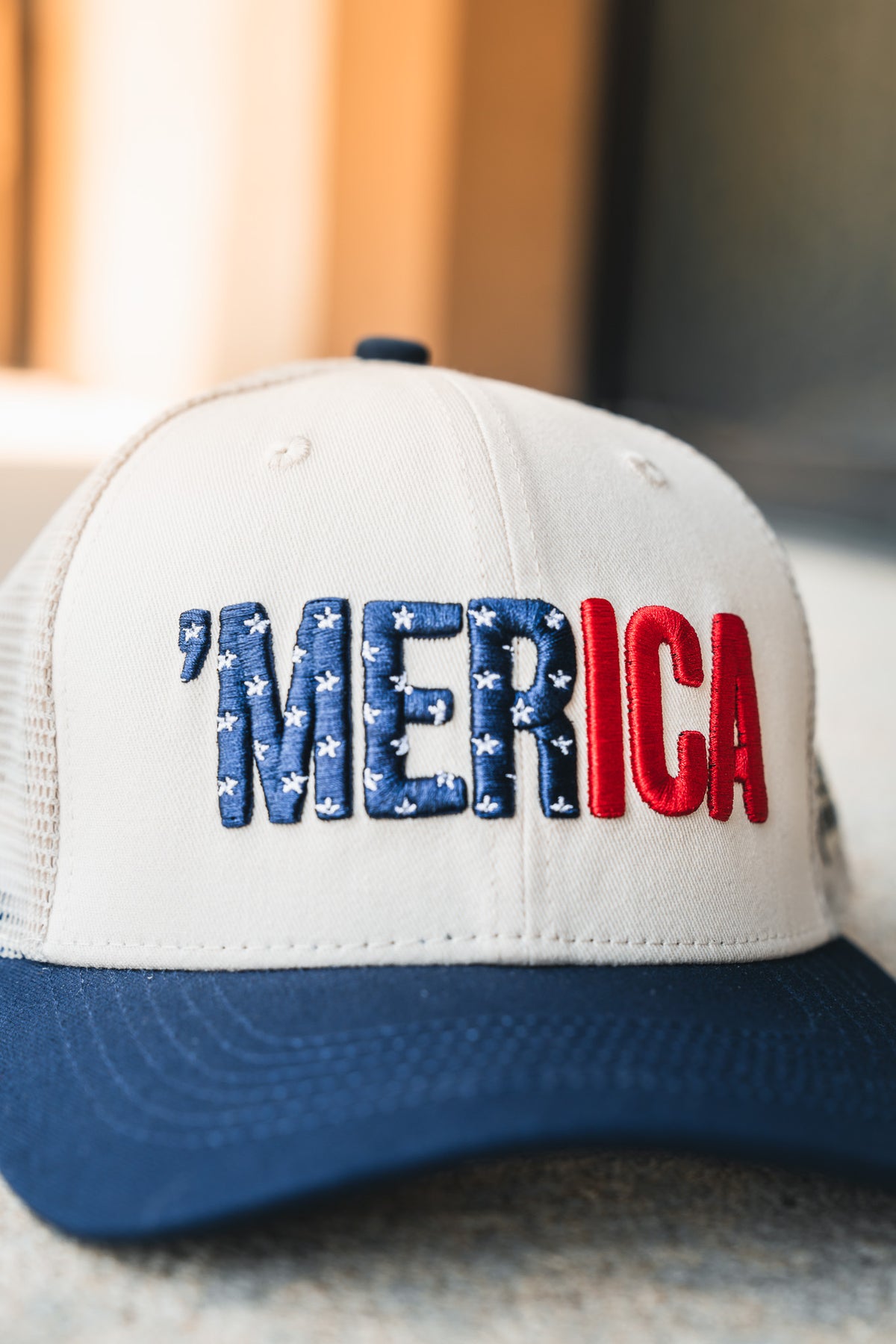 The Born United Private Label ’Merica Trucker Snapback is a beige and navy blue hat featuring “MERICA” embroidered on the front panel in blue with white stars and red letters.