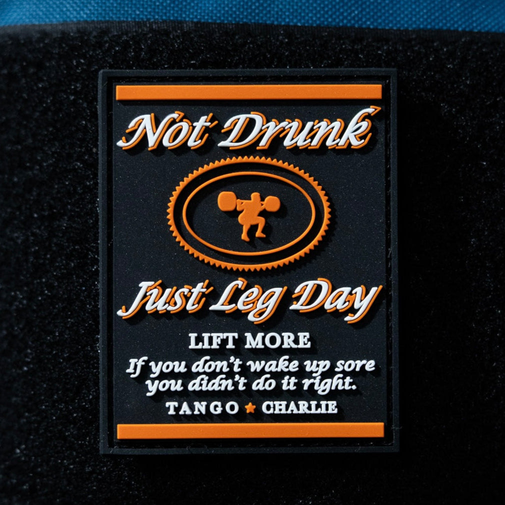 The Not Drunk, Just Leg Day - Patch by Tango Charlie Apparel is a 3" rectangular PVC patch featuring orange/white text, a weightlifter graphic, and motivational phrases like "LIFT MORE" and "If you don’t wake up sore you didn’t do it right.