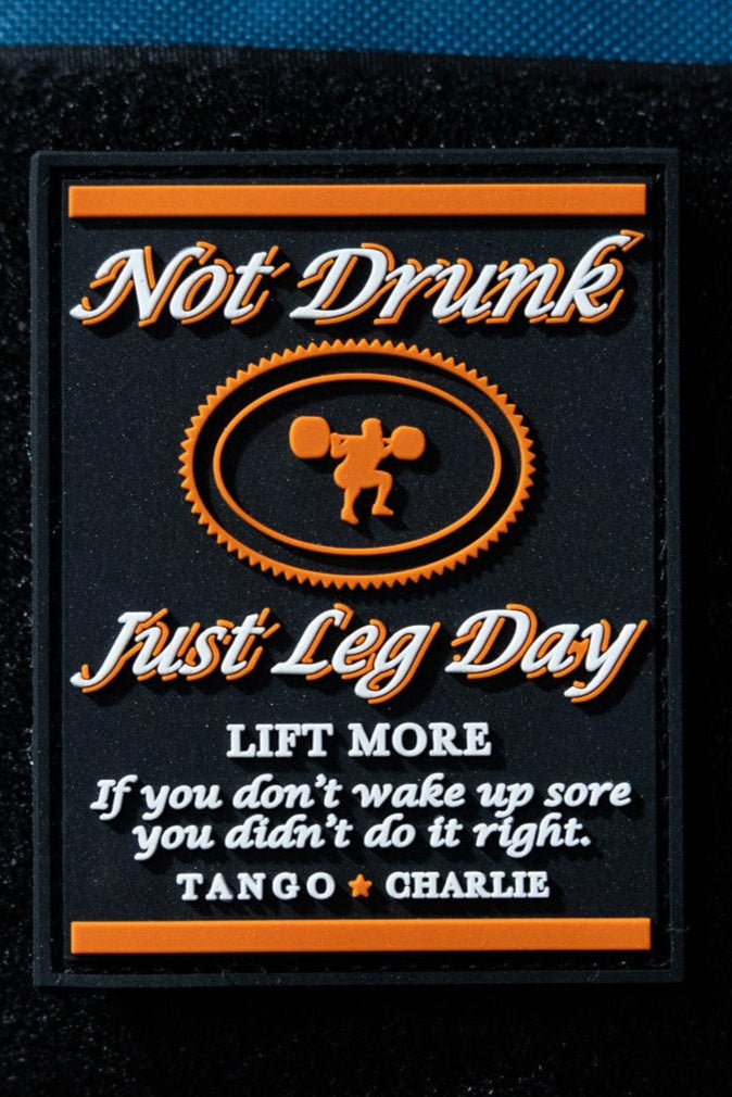 The Not Drunk, Just Leg Day - Patch by Tango Charlie Apparel is a 3" rectangular PVC patch featuring orange/white text, a weightlifter graphic, and motivational phrases like "LIFT MORE" and "If you don’t wake up sore you didn’t do it right.