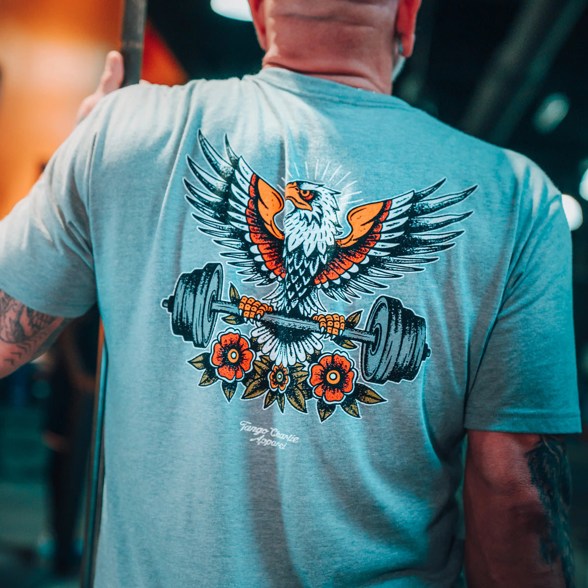 A tattooed person wears the Tango Charlie Apparel Old School Never Gets Old gray shirt with a colorful eagle graphic, holding Cold Iron weightlifting plates with flowers below, capturing an old school gym vibe.