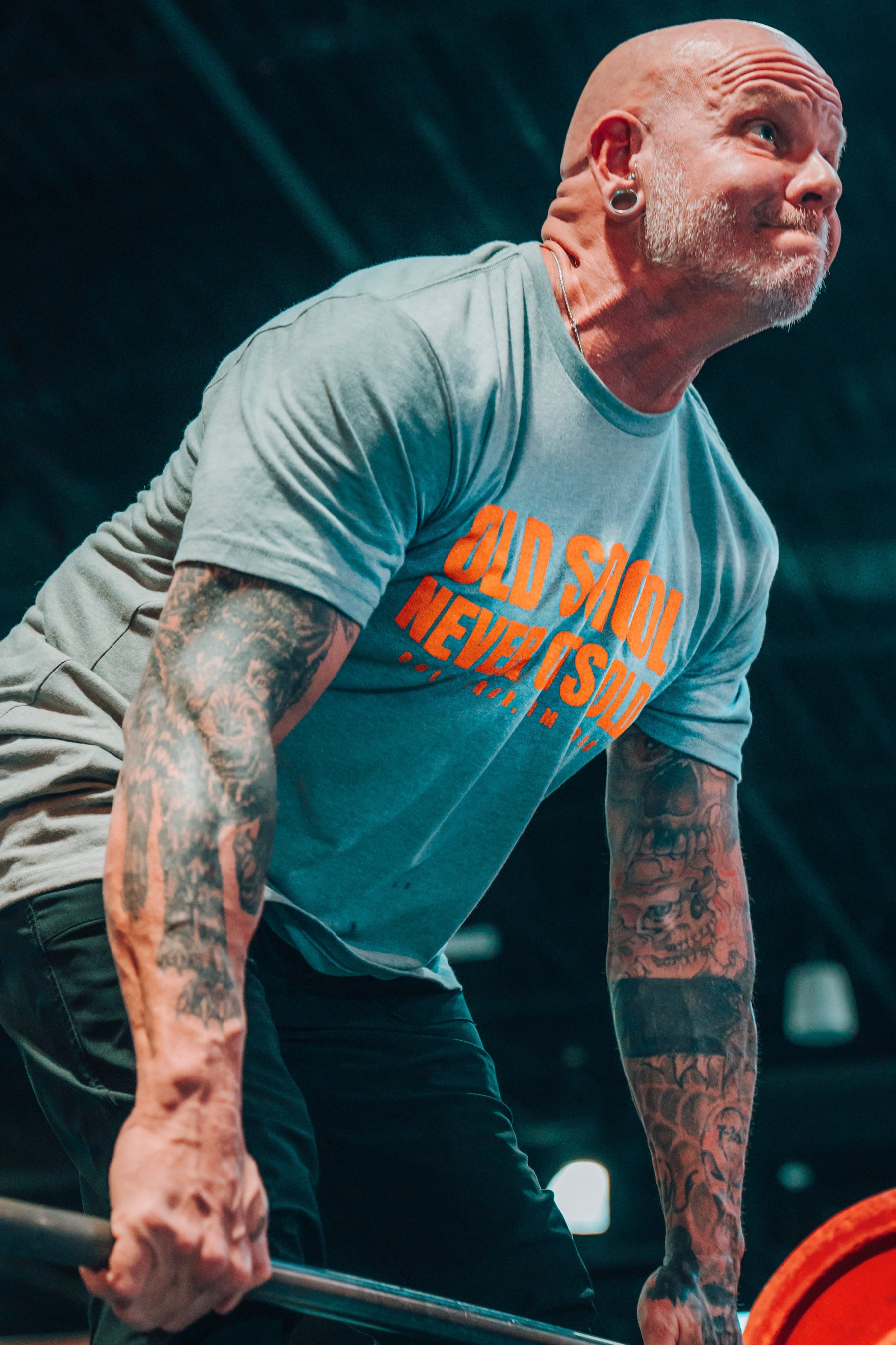 A muscular, tattooed man with a shaved head and large ear gauges lifts a barbell in an indoor gym, wearing Tango Charlie Apparel’s blue “Old School Never Gets Old” T-shirt. Classic weight plates are loaded on the bar.