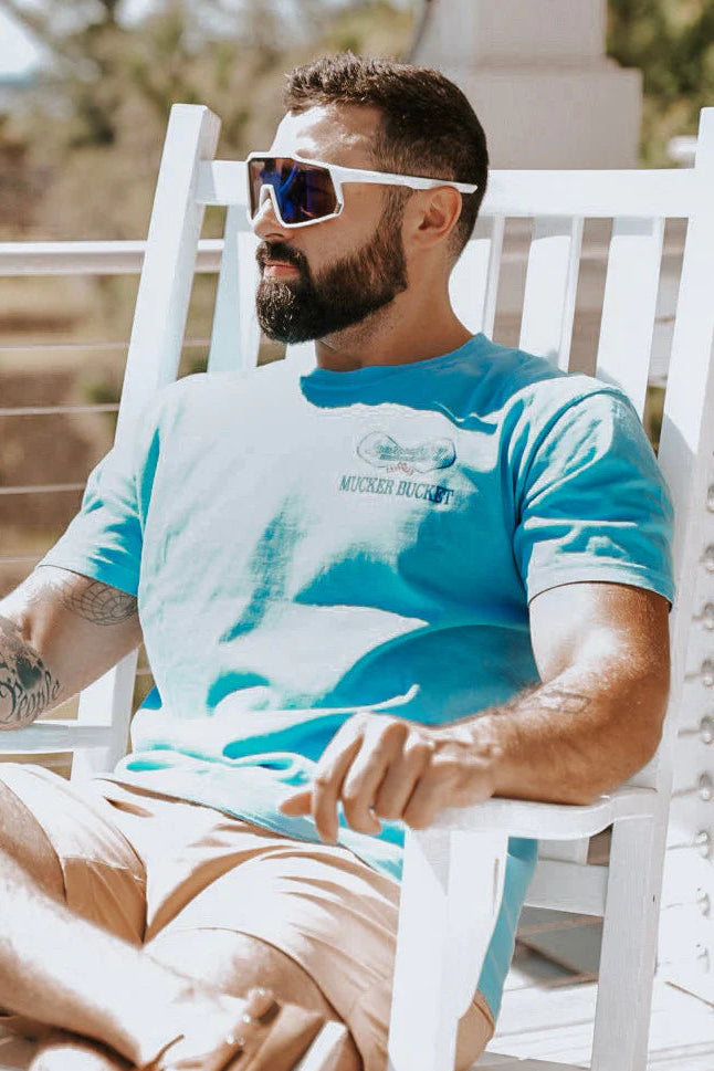 Man wearing a blue t-shirt with a logo, sunglasses, and beige shorts sitting on a white chair outdoors.