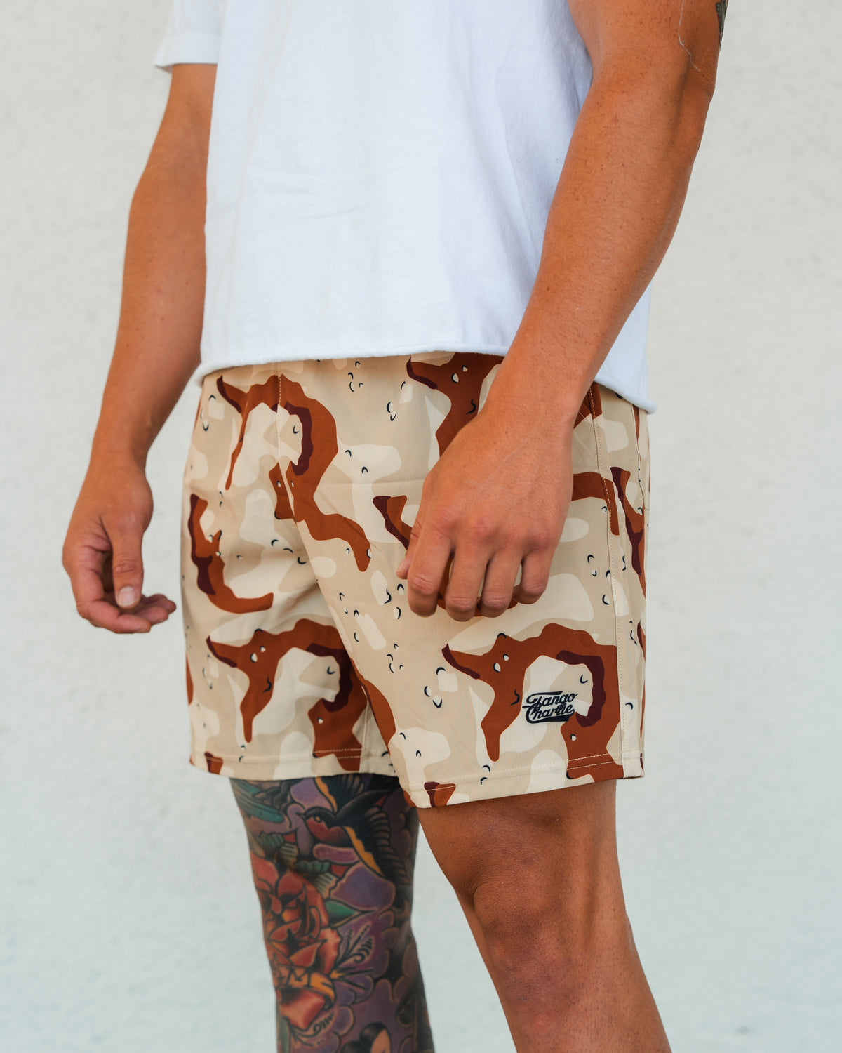 A person in a white T-shirt and Tango Charlie Apparel Men's Performance Training Shorts | 5.5" – Choco Chip stands against a plain light background. Their left leg has a colorful tattoo, with the shorts’ logo visible on the left leg.