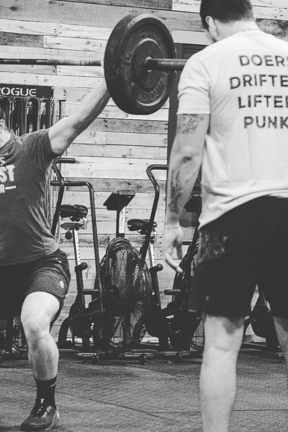 In a black and white gym scene, a man lifts a barbell overhead as another stands before him in the Tango Charlie Apparel Doers Drifters Lifters Punks Unisex Tee—perfect for showing off your crew’s spirit.
