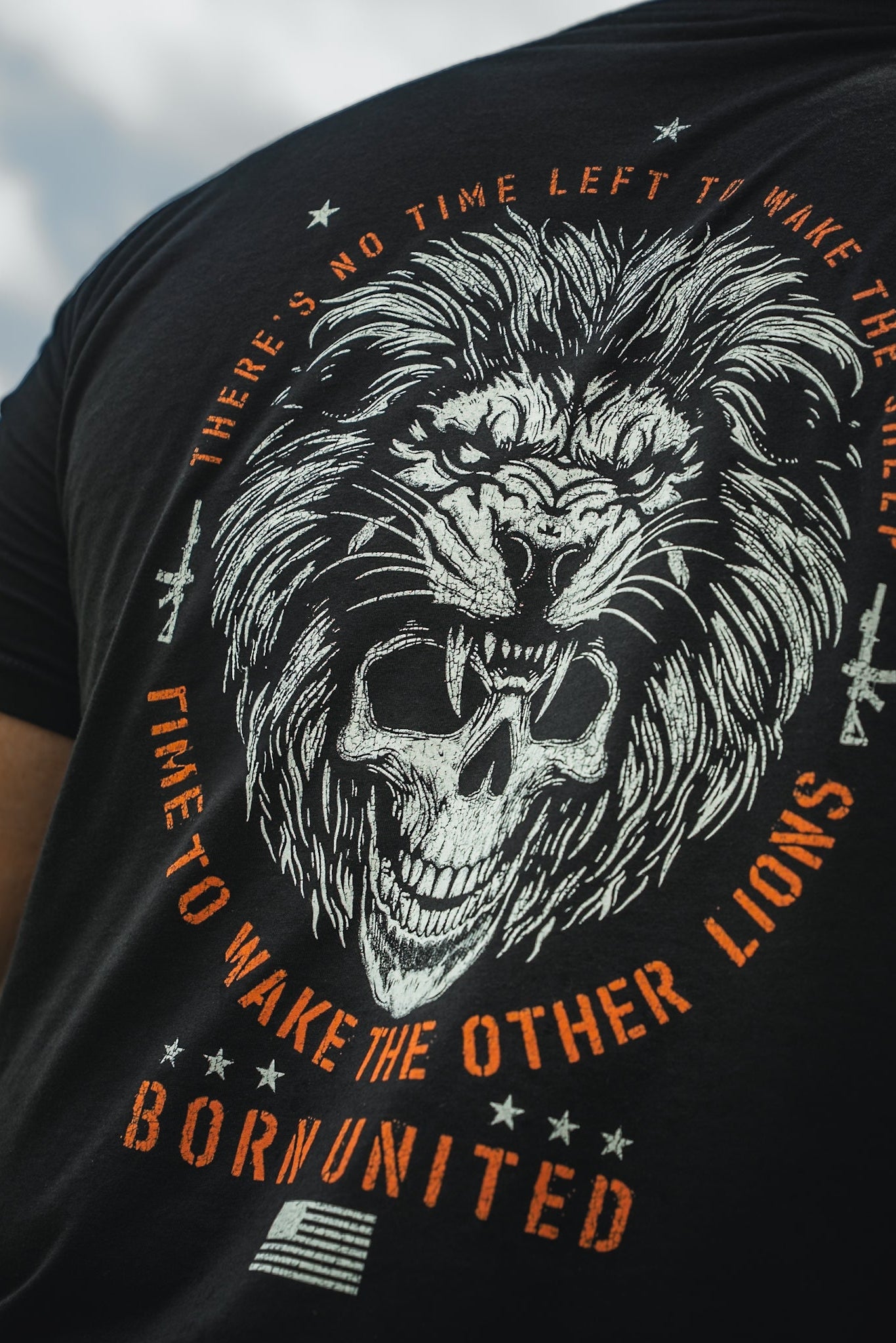 A person dons the Waking Lions T-shirt by Born United Private Label, featuring a black tee with a roaring lion graphic above a skull. The text reads, No time left to wake the sheep. Time to wake the other lions, and Born United is displayed above an American flag at the bottom.