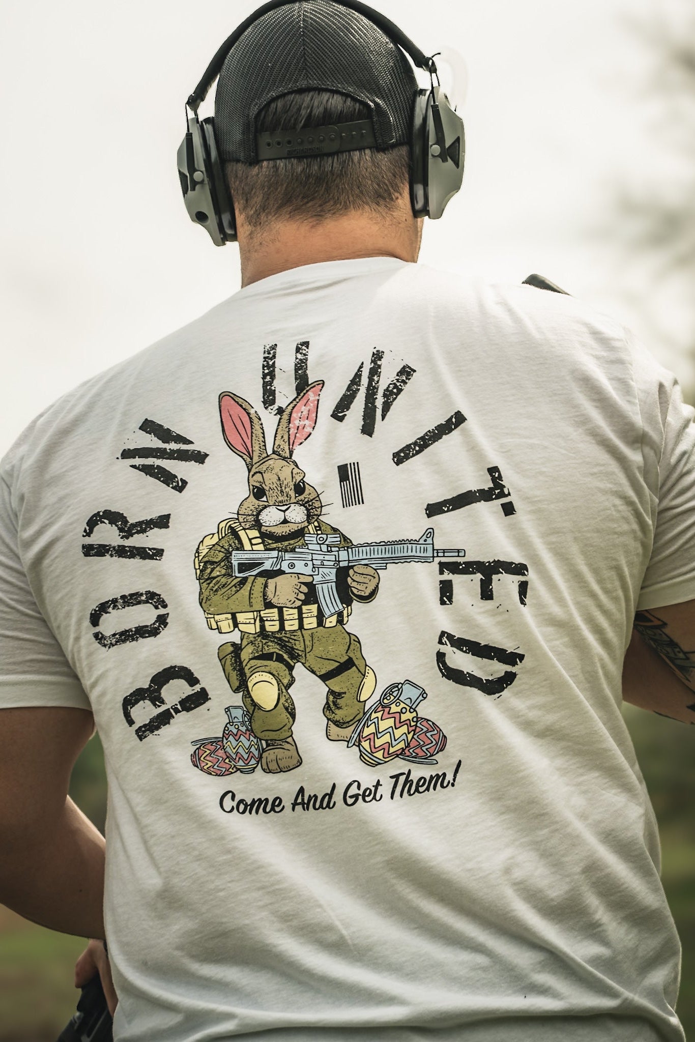 A person in headphones and a white "Battle Bunny" tee by Born United Private Label, featuring a bunny in military gear with a rifle and Easter eggs. The shirt reads BORN UNITED and Come And Get Them!—a bold Easter dinner conversation starter.