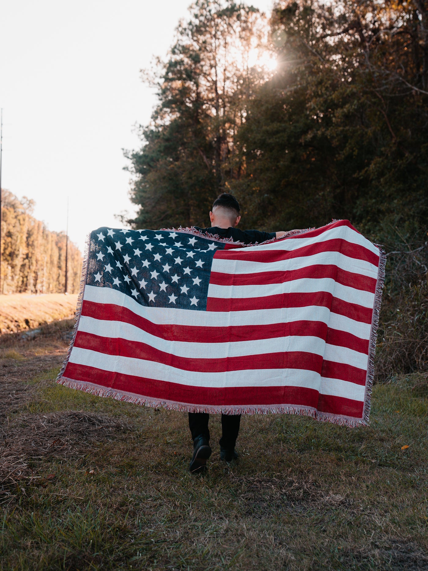 A person walks away on a grassy path, holding an American Flag Jacquard Blanket from Born United Private Label. The sun peeks through tall trees in the background, casting a warm glow on this quintessentially American scene.