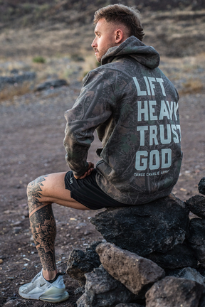 A man with leg tattoos sits on rocks outdoors, wearing the Lift Heavy. Trust God. - Premium Darkwood Camo Hoodie by Tango Charlie Apparel—a bold Christian fitness statement inspired by Proverbs 3:5-6.