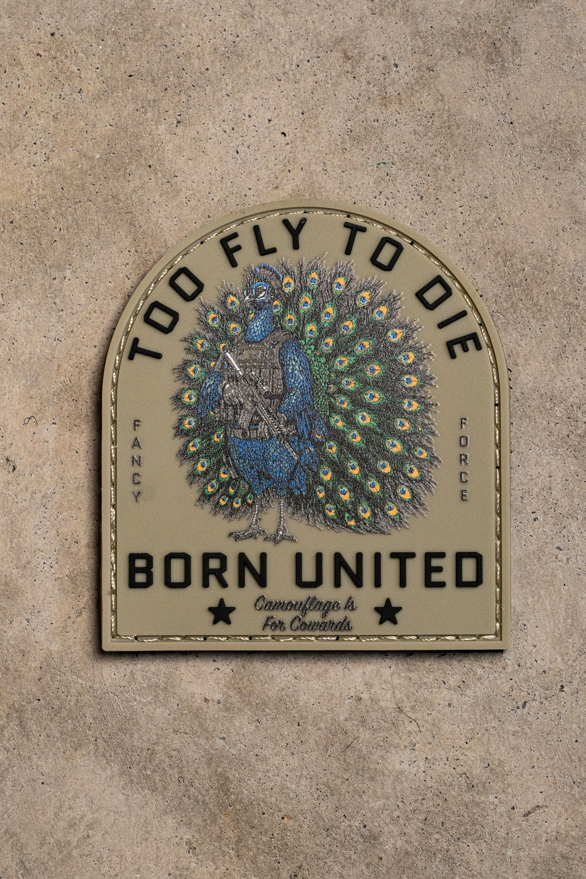 Tan arched 3D PVC Too Fly To Die patch featuring a peacock in body armor holding a rifle, with text reading: TOO FLY TO DIE, FANCY FORCE, BORN UNITED, and Camouflage is For Cowards.