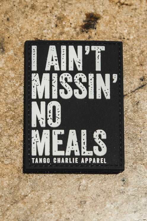 The I Ain’t Missin’ No Meals - Patch by Tango Charlie Apparel is a black 3-inch Velcro patch with bold white text and smaller branding, displayed on a textured brown surface.