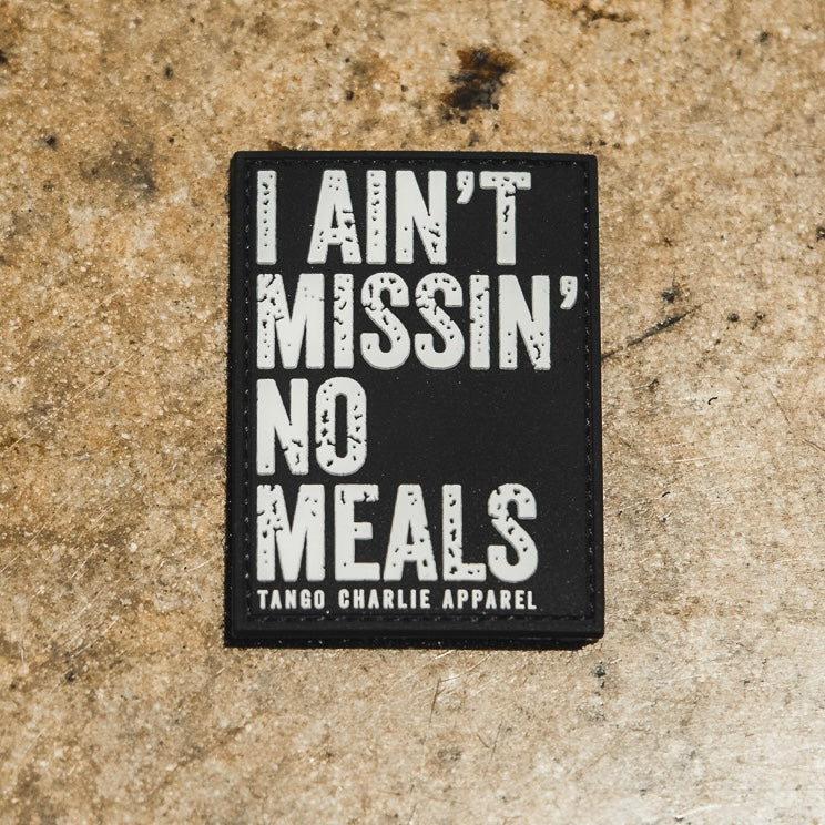 The I Ain’t Missin’ No Meals - Patch by Tango Charlie Apparel is a black 3-inch Velcro patch with bold white text and smaller branding, displayed on a textured brown surface.