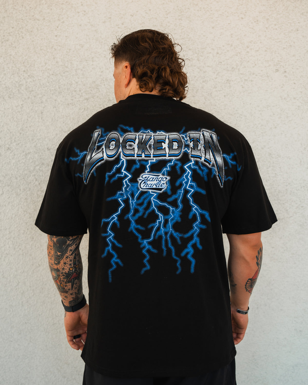 A tattooed individual faces away, wearing Tango Charlie Apparel’s Locked In - Premium Oversized Tee, featuring blue lightning graphics and “LOCKED IN” plus “Stand Chance” text printed on the back.