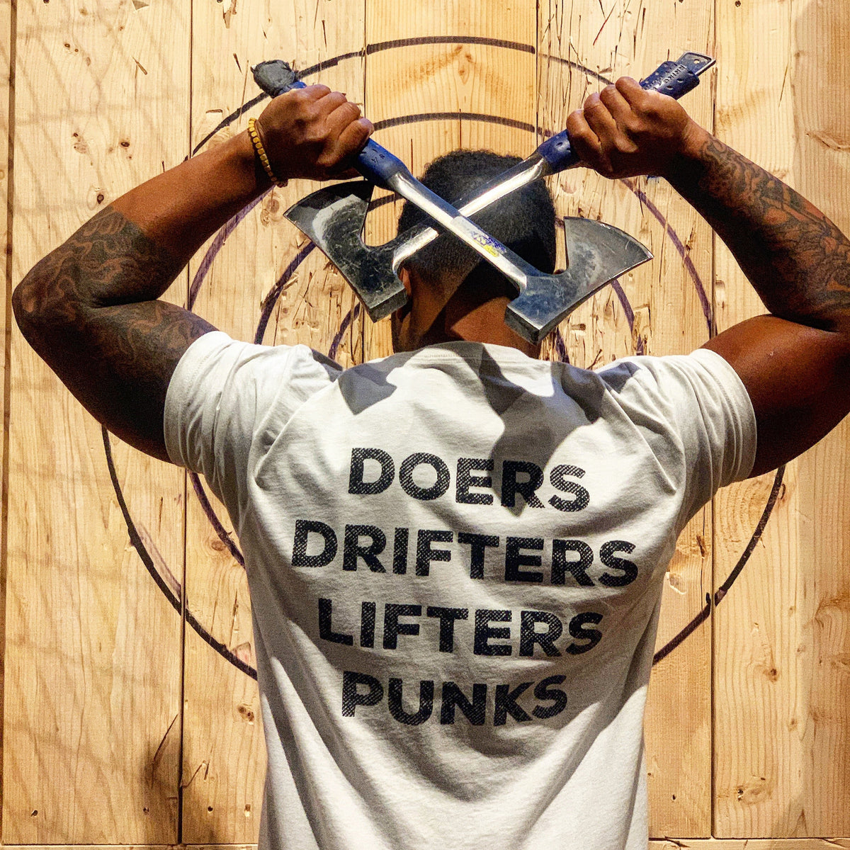 A person stands before a wooden target wall, holding two crossed axes behind their head and rocking the Tango Charlie Apparel Doers Drifters Lifters Punks Unisex Tee—perfect for repping your crew in style.