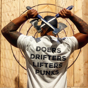 A person stands before a wooden target wall, holding two crossed axes behind their head and rocking the Tango Charlie Apparel Doers Drifters Lifters Punks Unisex Tee—perfect for repping your crew in style.