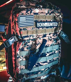 The Digi-Cam Tac-Pac - 45L from Born United Private Label, featuring American flag and Born United patches plus a sheathed black knife on the front, is illuminated by blue and red lights—ideal for carrying essentials or your digital camera.