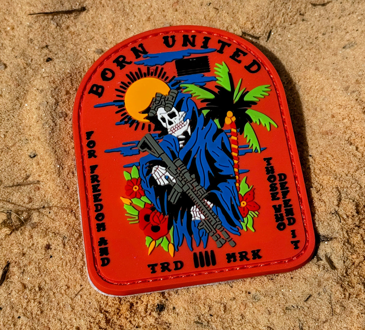 The Born United Always Deadly PVC Patch features a skeleton in a blue cloak holding a rifle on sand, with sun, palm trees, and roses. The text reads: BORN UNITED FOR FREEDOM AND THOSE WHO DEFEND IT TRD MRK.