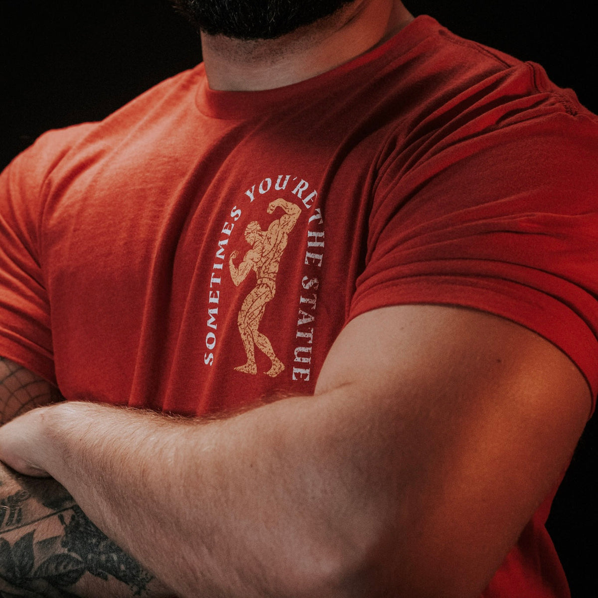 A person with crossed arms wears Tango Charlie Apparel’s “St. Arnold The Stoic” red t-shirt, featuring a bodybuilder illustration and the text “SOMETIMES YOU’RE THE STATUE.” Their face is not visible.