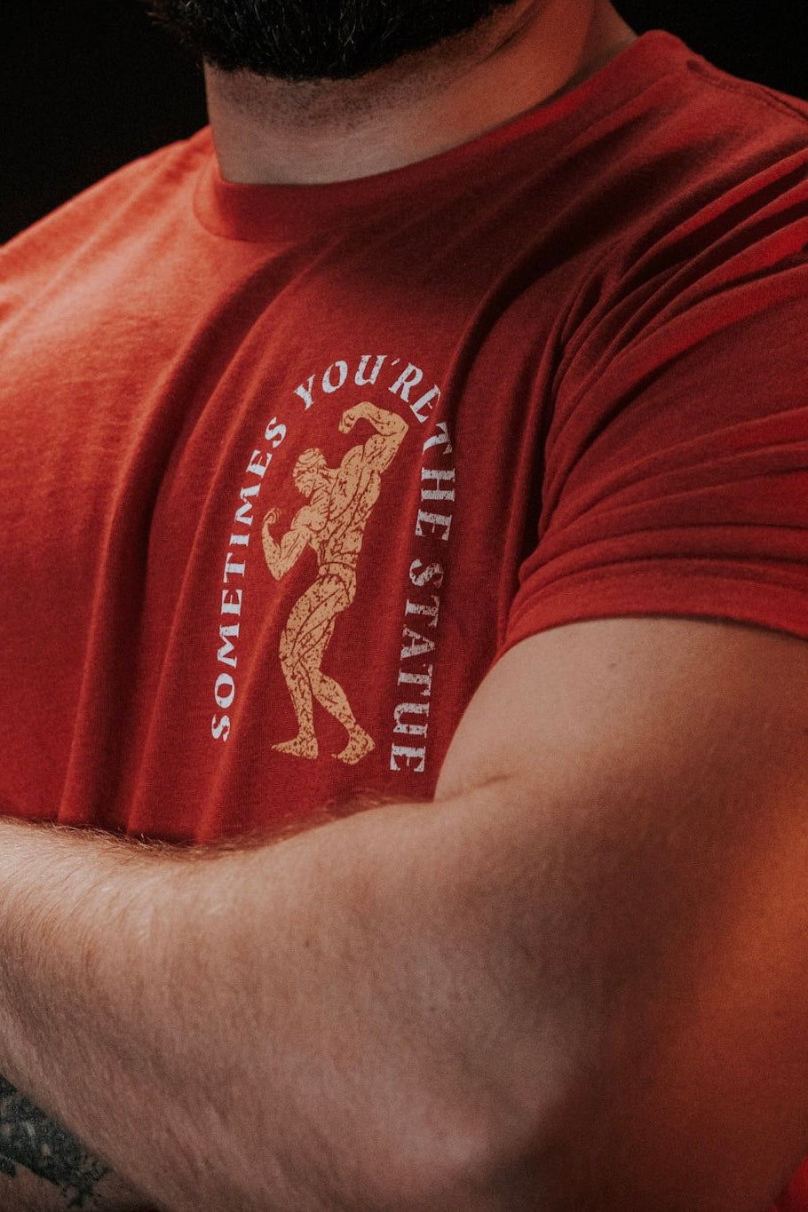 A person with crossed arms wears Tango Charlie Apparel’s “St. Arnold The Stoic” red t-shirt, featuring a bodybuilder illustration and the text “SOMETIMES YOU’RE THE STATUE.” Their face is not visible.