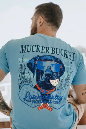 Man wearing a light blue t-shirt with 'Mucker Bucket' and 'Lowcountry' graphics, sitting outdoors.