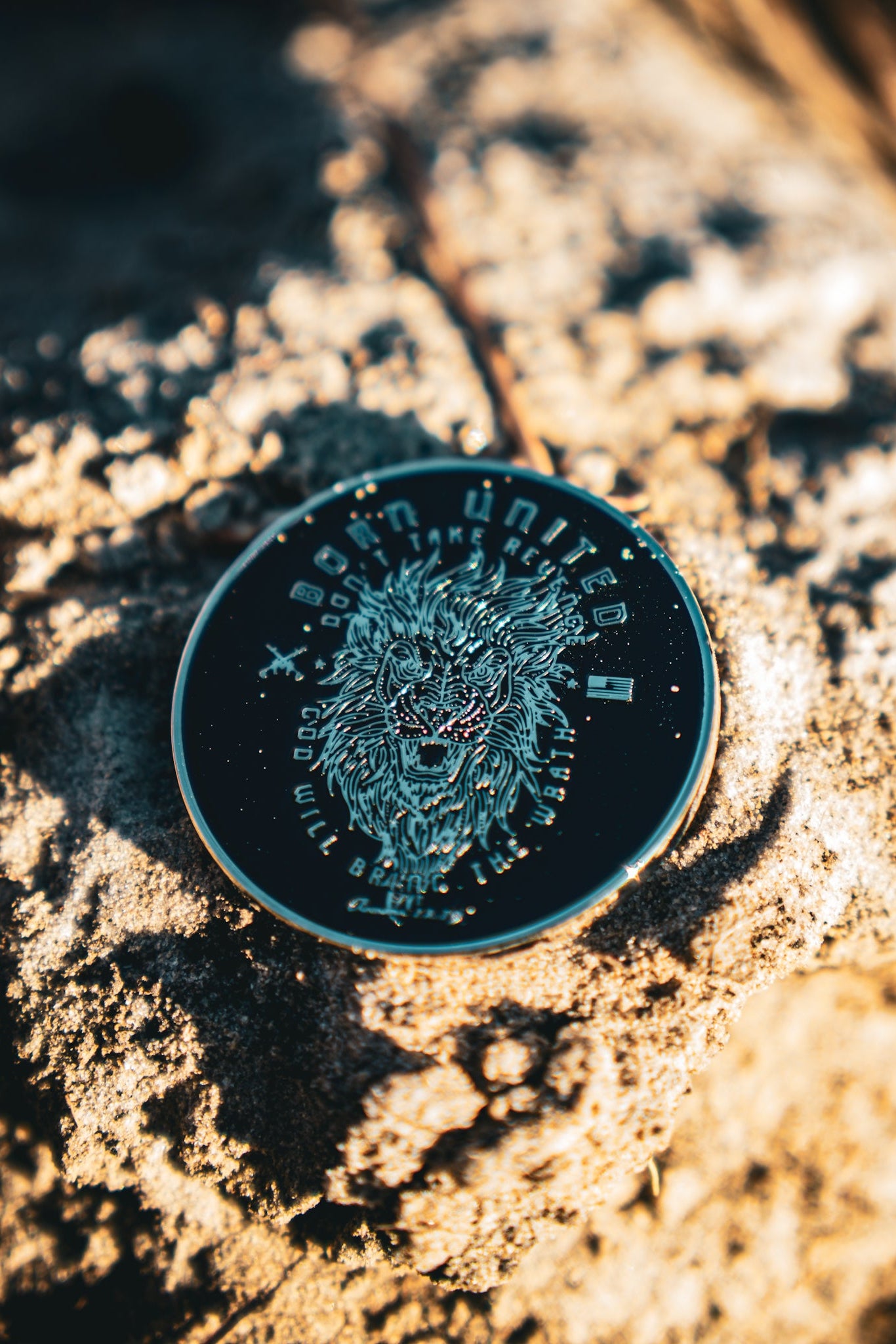 The Revenge Challenge Coin by Born United Private Label, featuring a roaring lion, rests on a textured stone surface. Its intricate engravings are beautifully illuminated by sunlight, creating striking shadows.