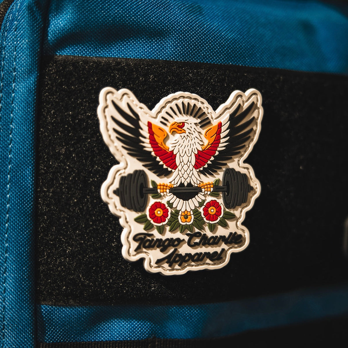 The "Old School, Never Gets Old - Patch" by Tango Charlie Apparel is a colorful 3D PVC patch showing an eagle with outstretched wings holding a barbell above two red flowers, attached to a blue fabric surface.