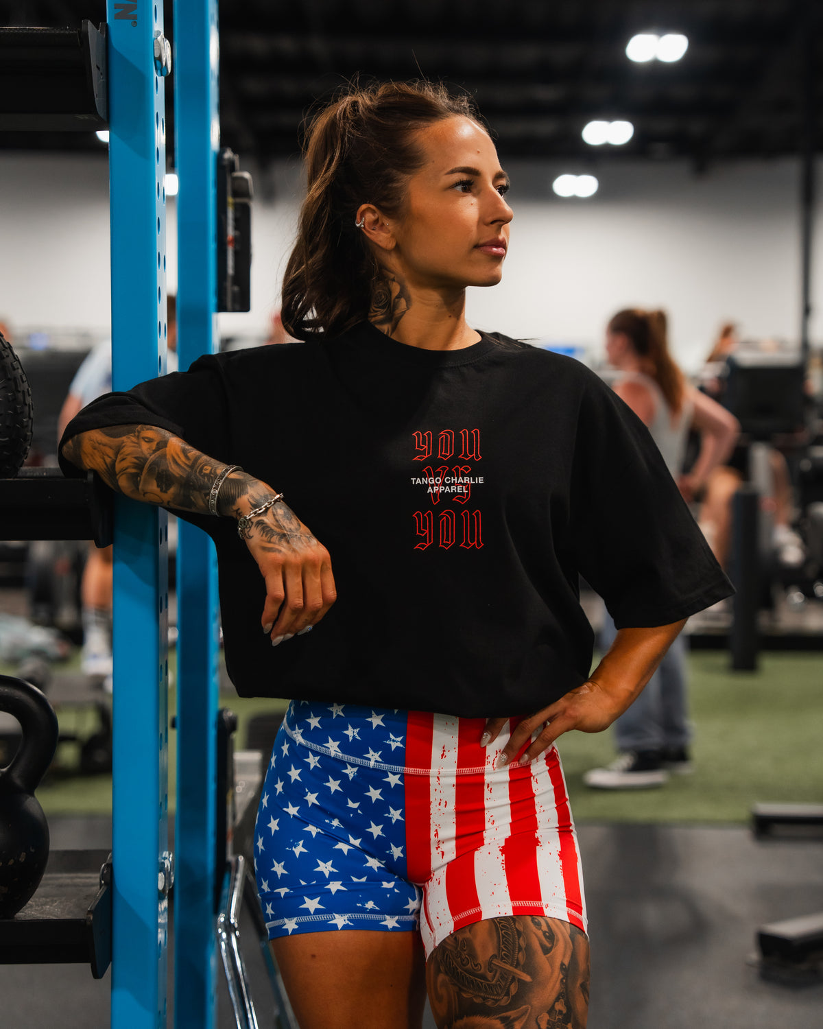 A tattooed woman stands in a gym, resting her arm on blue equipment. She wears a black T-shirt and Born United Private Label Women’s High Waisted Seamless Front Compression Training Shorts | Stars & Stripes. Other people and gym gear are visible.