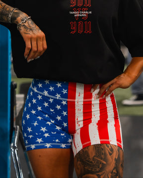 A tattooed woman stands in a gym, resting her arm on blue equipment. She wears a black T-shirt and Born United Private Label Women’s High Waisted Seamless Front Compression Training Shorts | Stars & Stripes. Other people and gym gear are visible.