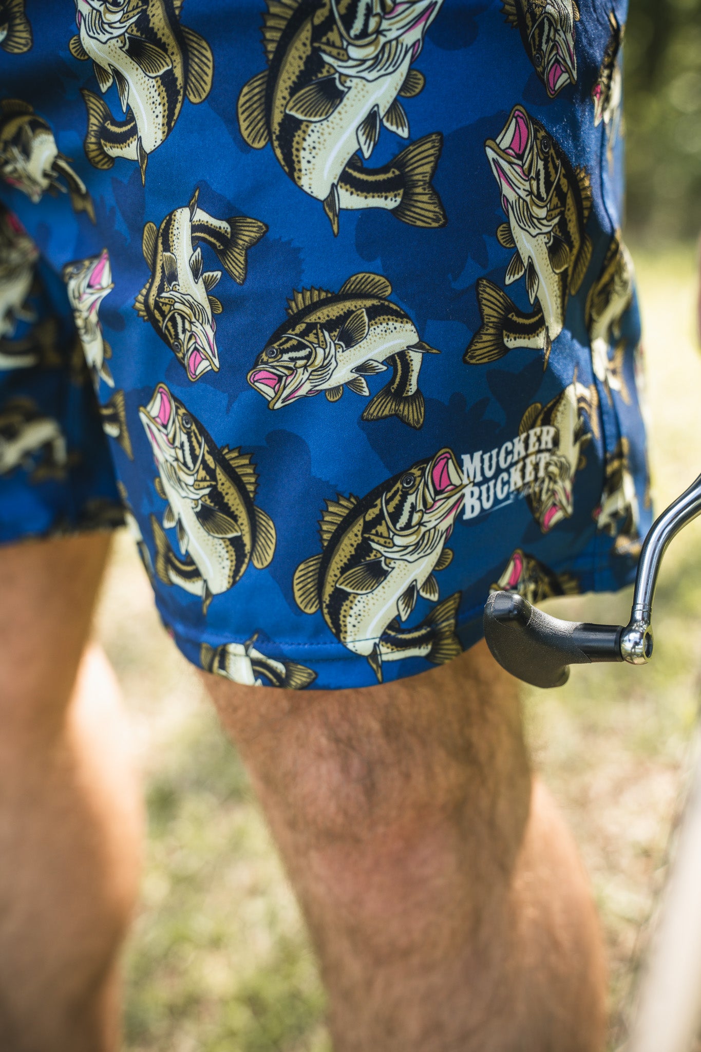 Close-up of a person’s leg wearing Mucker Bucket Men’s Performance Athletic Shorts | 5.5"–Big Bass Daddy with a fish pattern, near a bicycle pedal outdoors on grass.