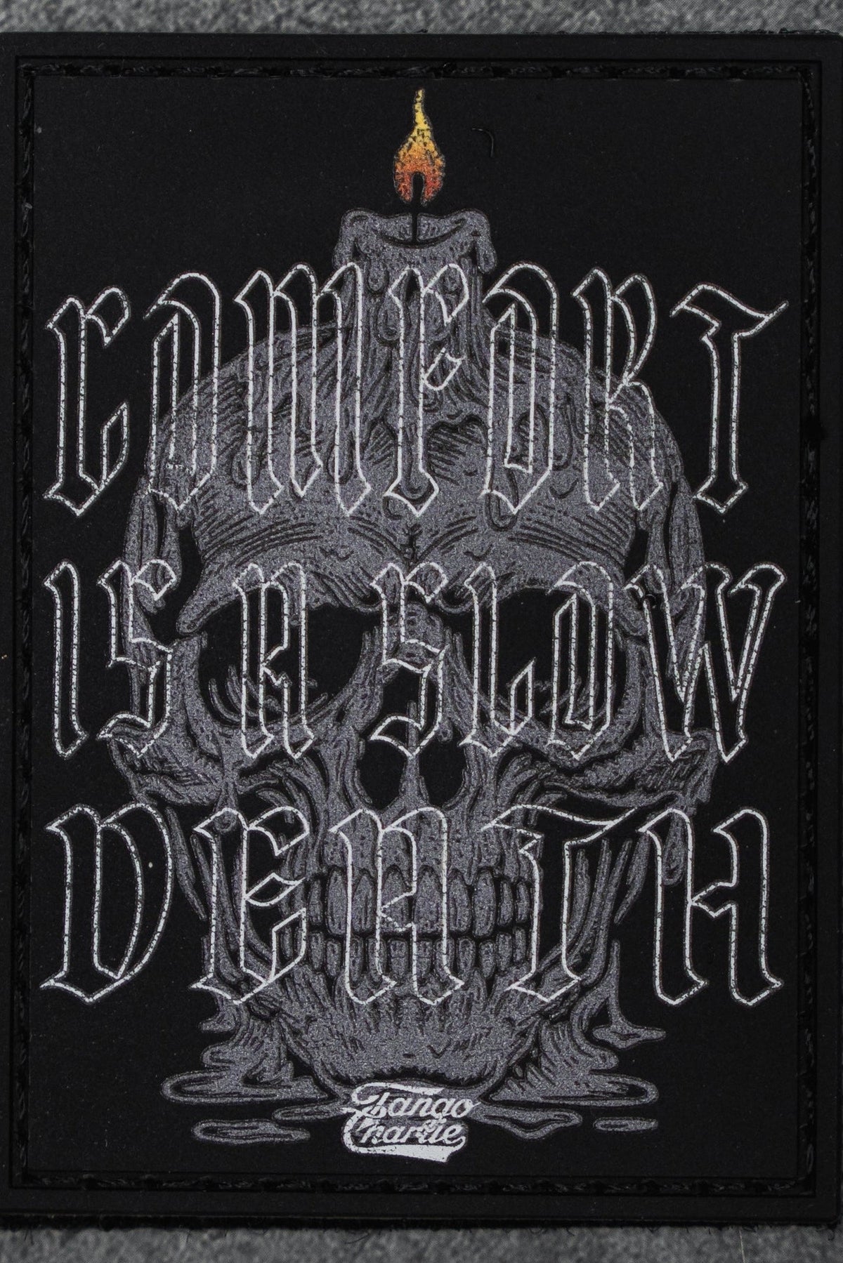 The Comfort is a Slow Death - Patch by Tango Charlie Apparel is a 3" Velcro patch with a gray skull and candle, featuring "COMFORT IS A SLOW DEATH" in stylized white letters and Slamco Charlie at the bottom.
