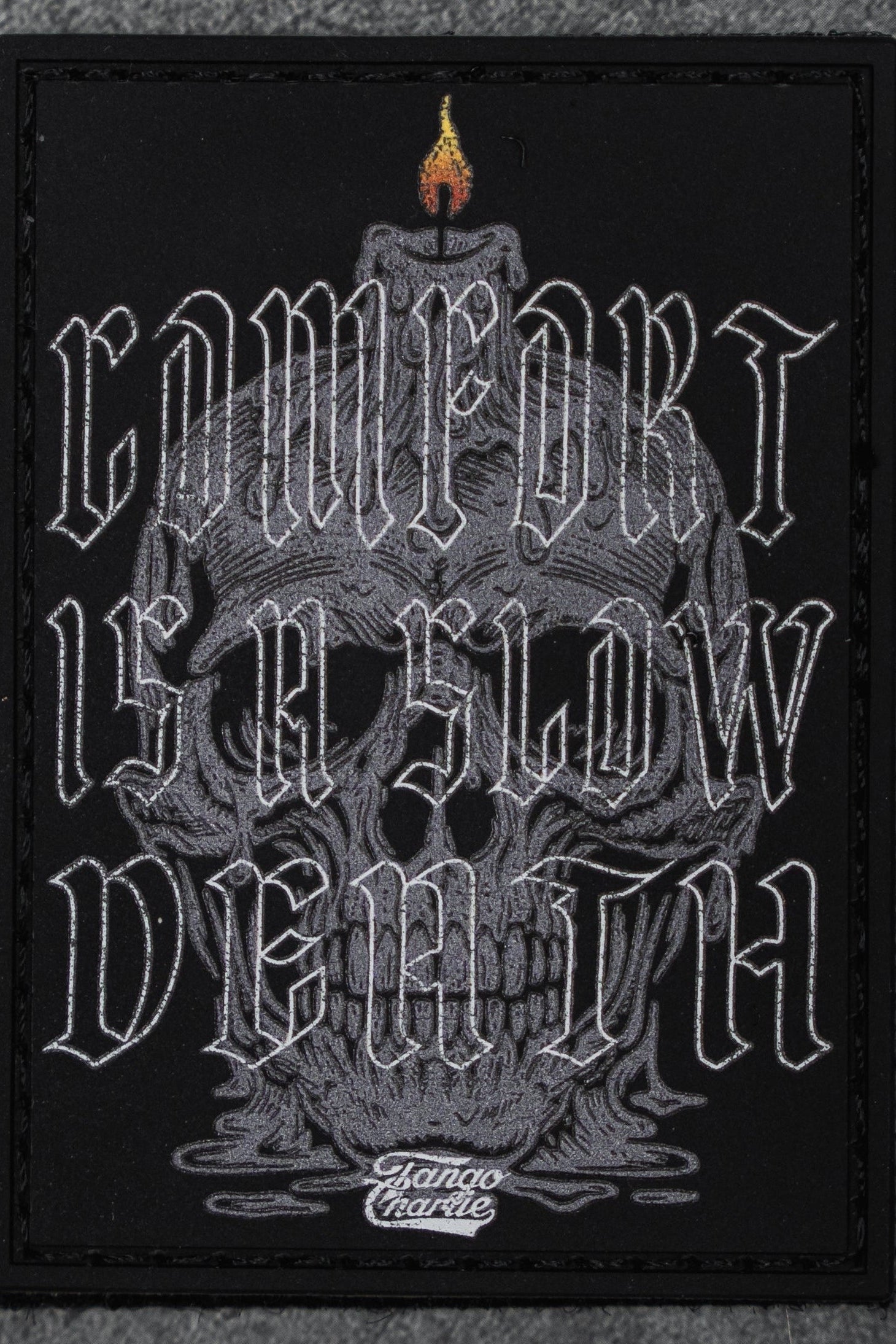 The Comfort is a Slow Death - Patch by Tango Charlie Apparel is a 3" Velcro patch with a gray skull and candle, featuring "COMFORT IS A SLOW DEATH" in stylized white letters and Slamco Charlie at the bottom.
