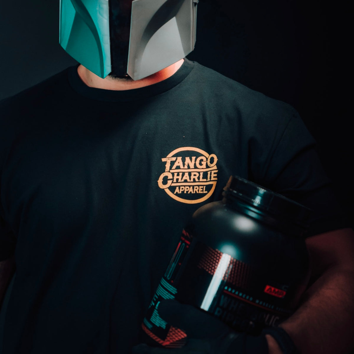 A person in a futuristic helmet and black Tango Charlie Apparel t-shirt holds a large container of The Whey protein against a dark background, ready for an epic workout.