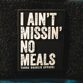 The I Ain’t Missin’ No Meals - Patch by Tango Charlie Apparel is a rectangular black 3D PVC patch with bold white text and the brand name below, displayed on textured black and blue fabric.