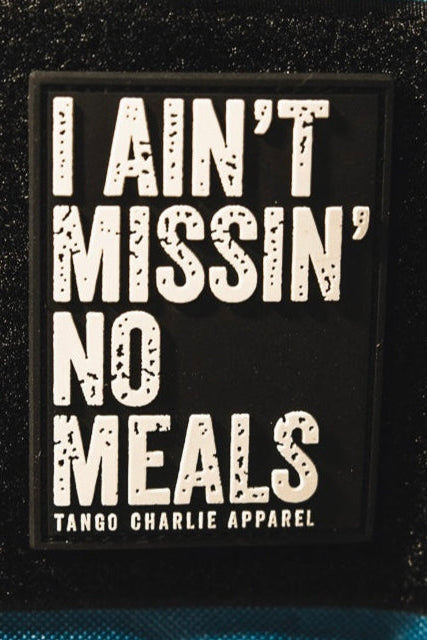 The I Ain’t Missin’ No Meals - Patch by Tango Charlie Apparel is a rectangular black 3D PVC patch with bold white text and the brand name below, displayed on textured black and blue fabric.