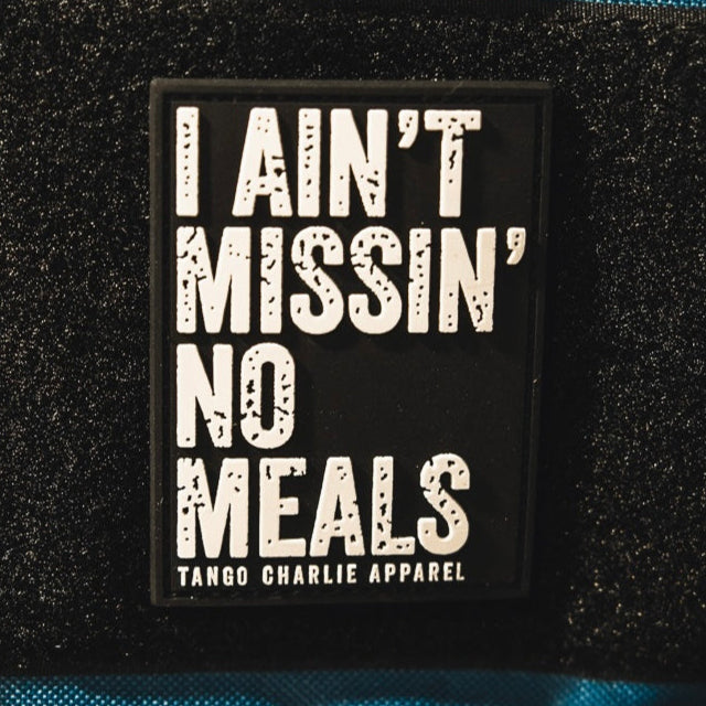 The I Ain’t Missin’ No Meals - Patch by Tango Charlie Apparel is a rectangular black 3D PVC patch with bold white text and the brand name below, displayed on textured black and blue fabric.