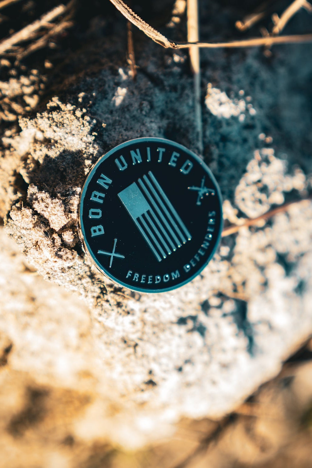 The Revenge Challenge Coin by Born United Private Label, resembling a round black badge with an abstract design and BORN UNITED, sits on textured rock. Sunlight and shadows from nearby dried grass enhance this rustic, natural setting.