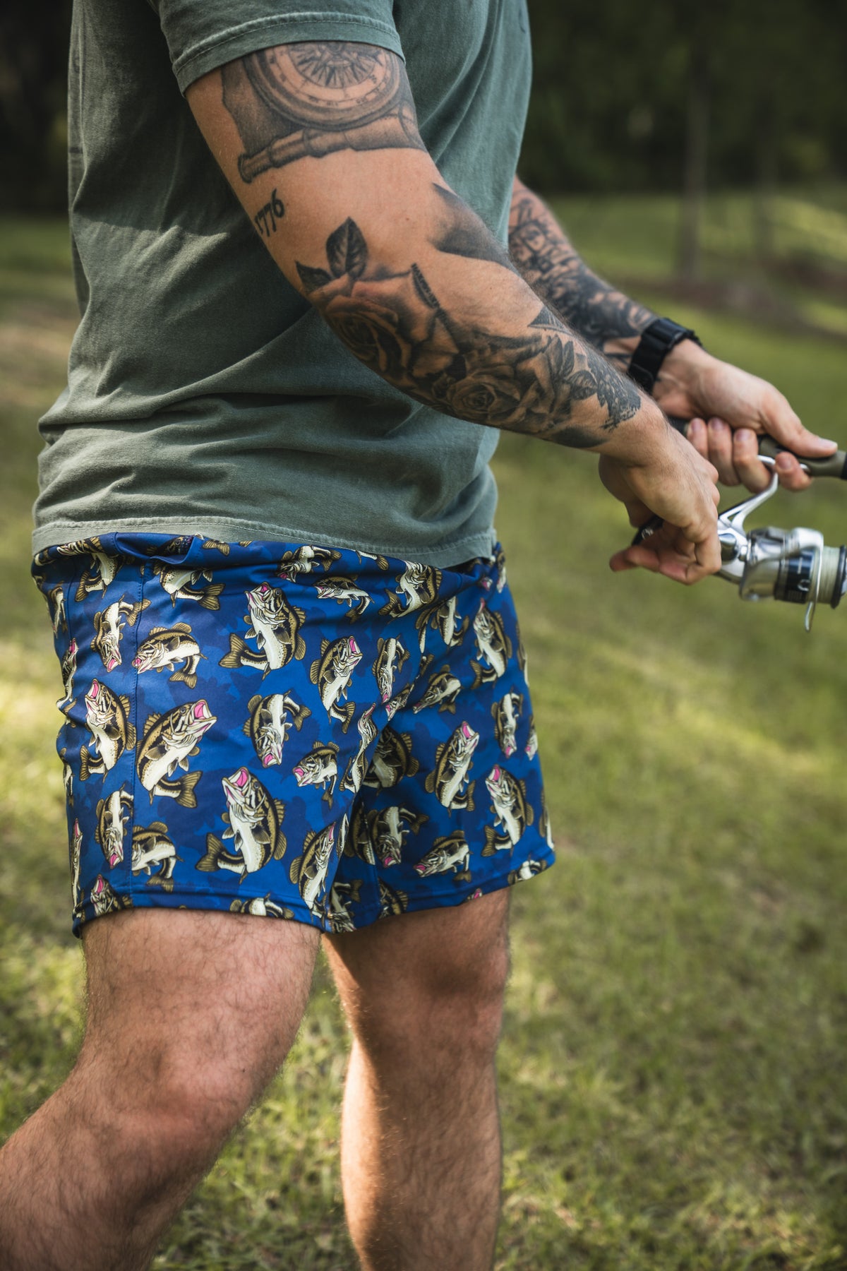 A tattooed person in a green shirt and Mucker Bucket Men's Performance Athletic Shorts | 5.5" - Big Bass Daddy with fish patterns stands on grass, holding a fishing rod.
